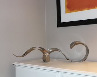May include: A silver metal sculpture of a wave, abstractly shaped, on a white dresser.