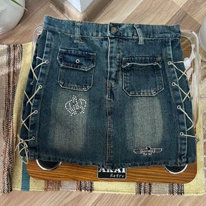 Op de afbeelding: Een blauwe denim rok met witte veters aan de zijkanten. De rok heeft twee voorzakken en een klein patch aan de voorkant met strass. De rok heeft een klein patch aan de achterkant met de tekst "AIR FORCE 24/7".