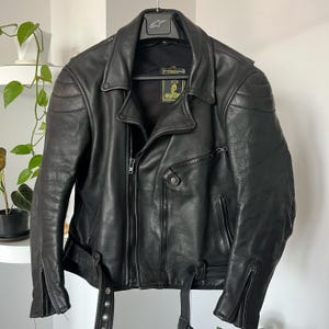 Op de afbeelding: Een zwart leren motorjack met een ritssluiting en een drukknoopsluiting. De jas heeft een 'GUCCI'-label aan de binnenkant.