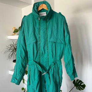 May include: Teal vintage ski suit with a high collar and multiple zips. The one-piece suit features a belted waist, shoulder epaulettes, and the brand name "stenebronn" on the collar. The suit is made of a textured material.