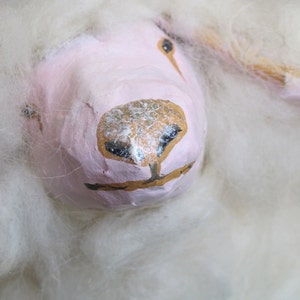 May include: A white fluffy sheep with a pink face and brown painted details. The sheep's face is painted with a brown nose and black eyes.