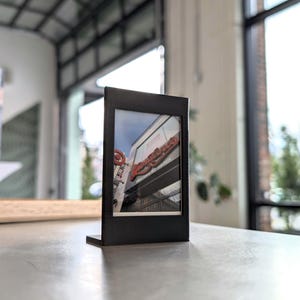 May include: A black picture frame with a built-in stand displays a photograph of a business sign. The sign reads "Longo's" in red lettering. The frame is on a light grey surface.