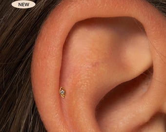 Può includere: Primo piano di un orecchio con un piccolo orecchino dorato. L'orecchino ha una forma a goccia con una piccola gemma trasparente al centro. La parola "NEW" è in un ovale bianco in alto a sinistra.