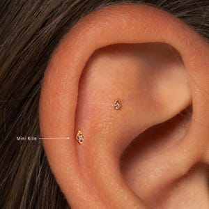 May include: Close-up of an ear with two small, gold-colored earrings. One earring is kite-shaped, the other is teardrop-shaped. Both earrings have a small, clear gem in the center. The text "Mini Kite" is next to the kite-shaped earring.
