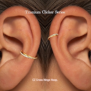 May include: Close-up of two ears each adorned with a gold CZ Cross Hinge Hoop earring. The earrings are embellished with small, sparkling stones. The text "Titanium Clicker Series" is visible at the top of the image.