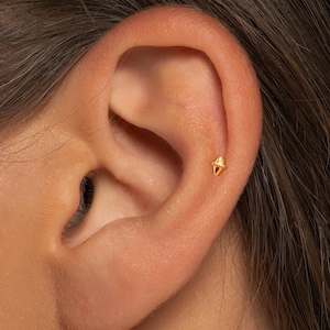 May include: Close-up of a gold stud earring with a geometric design. The small earring is set on the upper ear cartilage. The earring has a pointed shape.