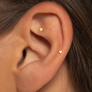 May include: Close-up of an ear with two small, gold heart-shaped stud earrings. The earrings are positioned on the upper ear and the earlobe. The skin tone is light, and the earrings provide a subtle, elegant accent.