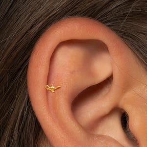 May include: A small, gold-colored earring in the shape of a genie lamp is shown. The earring is placed on the upper ear cartilage. The background is a close-up of a person's ear and hair.