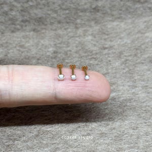 以下が含まれることがあります： 小さな白いオパールストーンが付いたゴールドのボディジュエリー4点。ジュエリーは指に飾られています。