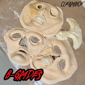 May include: Three beige foam prosthetic face masks with skull-like features, including eye sockets, nasal openings, and exposed teeth. The text CLEARANCE is in the top right and B-GRADES is in the bottom left of the image.