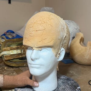 May include: A white foam mannequin head with a tan, textured material applied to the top. The head is on a patterned, gray surface. A hand wearing a brown glove is visible.