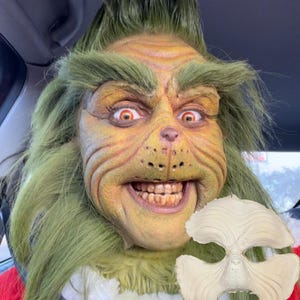 Puede incluir: Primer plano de una máscara y un disfraz del Grinch. La máscara es verde y amarilla con ojos rojos y una amplia sonrisa. También se ve una máscara separada sin pintar. El disfraz incluye piel verde y un atuendo rojo.