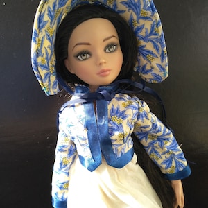 Peut inclure: Une poupée aux longs cheveux noirs portant une robe blanche avec une veste à imprimé floral bleu et jaune et un bonnet à imprimé floral bleu et jaune.