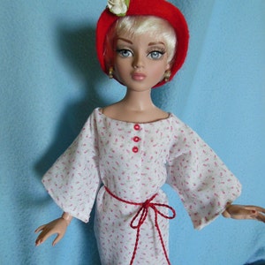 Puede incluir: Una muñeca con un vestido blanco con estampado floral rojo y un sombrero rojo con una flor blanca. La muñeca tiene el pelo rubio y los ojos azules.