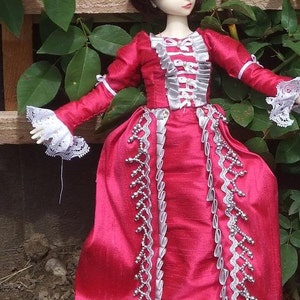 May include: A doll wearing a red dress with silver trim and white lace accents. The dress has a ruffled bodice and a long skirt. The doll has dark brown hair and closed eyes.