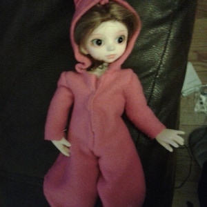 May include: A pink bunny rabbit costume for a doll. The costume has a hood with bunny ears and a zipper down the front. The doll has brown hair and blue eyes.