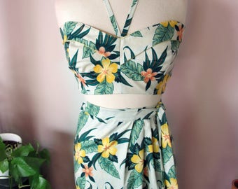May include: A white halter top and matching skirt with a tropical floral print featuring green leaves and yellow flowers. The top has a sweetheart neckline and the skirt is a wrap style.