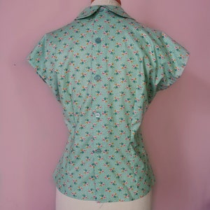 May include: A light green short-sleeved blouse with a white collar and a floral print. The blouse has a button closure in the back.