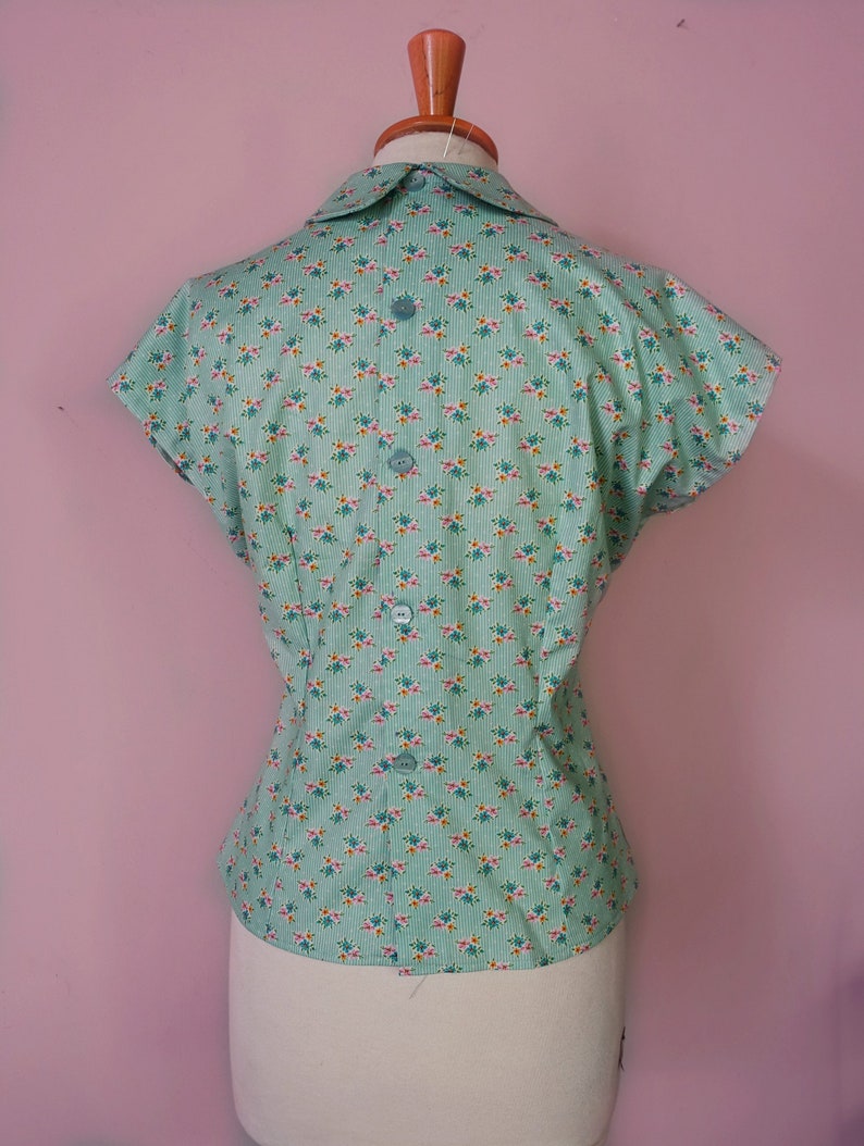 May include: A light green short-sleeved blouse with a white collar and a floral print. The blouse has a button closure in the back.