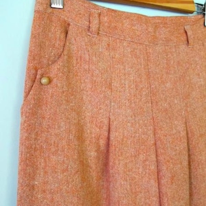 Könnte beinhalten: Nahaufnahme einer orangefarbenen Tweedhose, die an einem Holzbügel hängt. Die Hose hat eine geknöpfte Tasche und Falten. Der Stoff hat ein strukturiertes Aussehen.