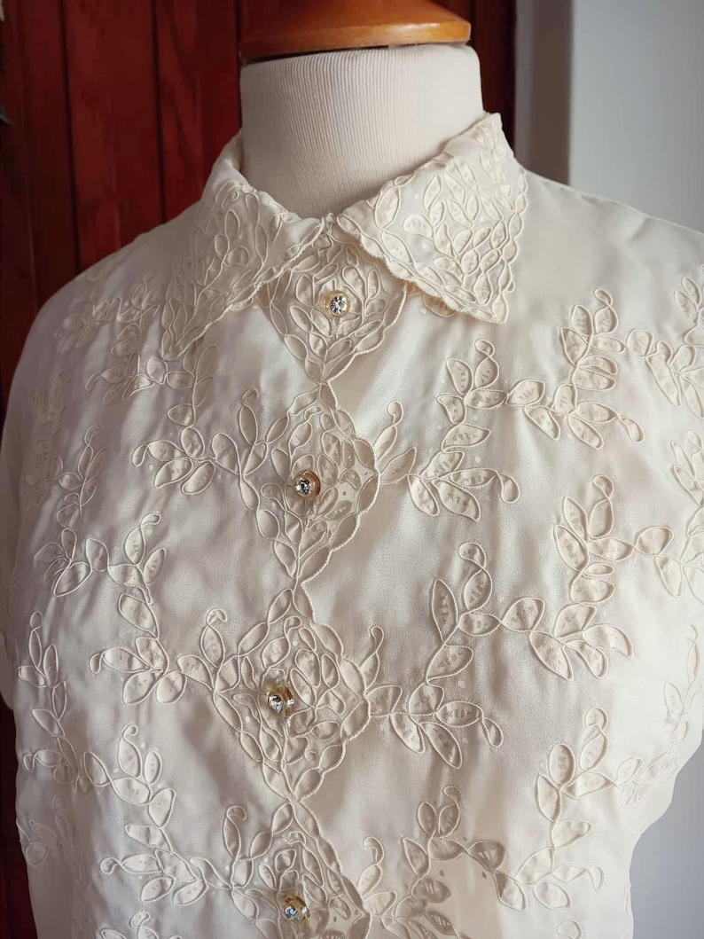May include: Ivory-colored blouse with intricate floral embroidery. The blouse features a classic collar and is adorned with decorative buttons. The delicate embroidery creates a textured effect, enhancing the overall elegance of the garment. The blouse is displayed on a mannequin.