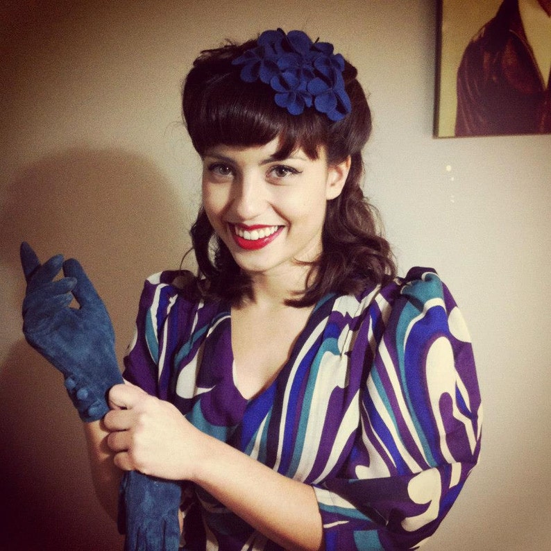 Puede incluir: Una mujer con cabello oscuro que lleva un vestido estampado en azul y blanco con un accesorio para el cabello de flores azules. Lleva guantes azules y tiene l&aacute;piz labial rojo.
