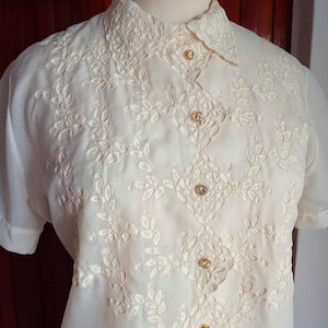 May include: Cream-colored short-sleeved blouse with intricate embroidered floral patterns. The blouse features a collared neckline and is fastened with decorative gold-toned buttons. The embroidery is detailed and covers the front and collar.