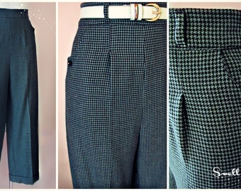 May include: High-waisted, wide-leg trousers in a black and teal houndstooth pattern. The pants feature pleats at the waist, side pockets with small black buttons, and a white belt with a gold buckle. The cuffs are turned up.