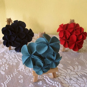 Puede incluir: Tres arreglos de flores de fieltro expuestos en peque&ntilde;os caballetes de madera. Las flores est&aacute;n hechas de fieltro y son de tonos azul, rojo y negro.