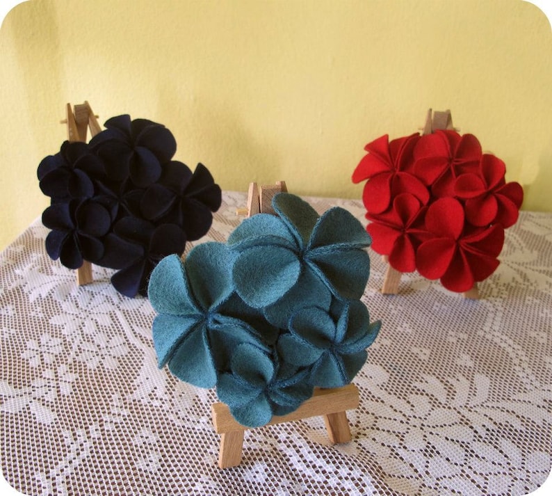 Puede incluir: Tres arreglos de flores de fieltro expuestos en peque&ntilde;os caballetes de madera. Las flores est&aacute;n hechas de fieltro y son de tonos azul, rojo y negro.