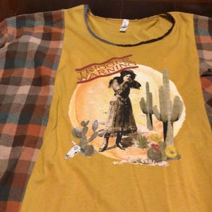 Puede incluir: Una camiseta de color amarillo mostaza con mangas de cuadros y un estampado gráfico que muestra a una persona en un paisaje desértico con cactus, una calavera y el texto TRIGGER WARNING y WILD LUCILLE. Cuello redondo con borde oscuro.
