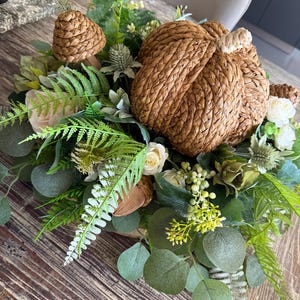 May include: A decorative arrangement featuring a large, woven brown pumpkin made of braided material, surrounded by artificial greenery including fern fronds, eucalyptus leaves, small white roses, thistle flowers, and a woven mushroom figure.