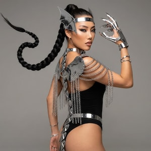 Puede incluir: Un disfraz de fantasía plateado con una diadema con detalles de alas, un collarín, una armadura para los hombros con detalles de cadena y guantes a juego. El disfraz se combina con un body negro. La modelo tiene una larga trenza.