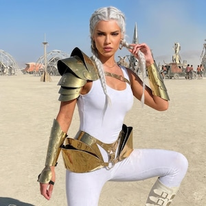 May include: A person wearing a white bodysuit and gold-colored armor, including shoulder pads, arm guards, and a waist belt. The armor has a metallic finish. The background shows a desert landscape with structures and people.