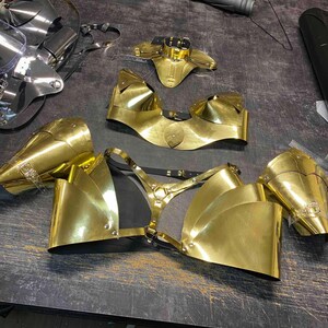 May include: A set of gold-colored metal body armor pieces, including a chest piece, shoulder pieces, and a neck piece. The pieces are made of metal and have a shiny finish.