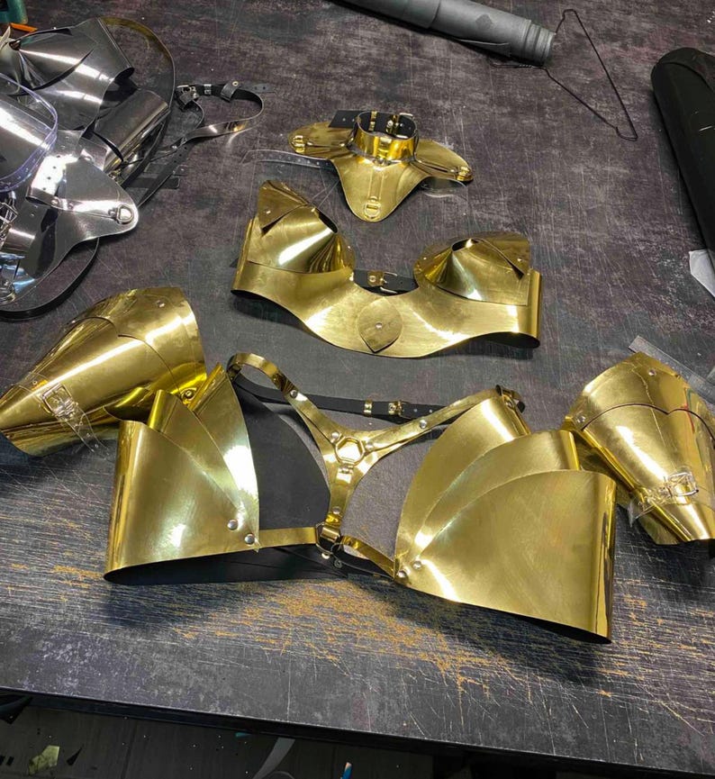 May include: A set of gold-colored metal body armor pieces, including a chest piece, shoulder pieces, and a neck piece. The pieces are made of metal and have a shiny finish.