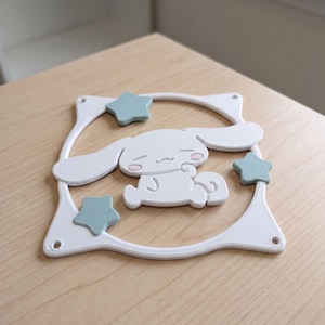 May include: A white square computer fan grill featuring a central cartoon character with long ears, closed eyes, and pink cheeks. The design includes three light blue stars positioned around the character, all set within a white frame.