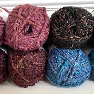 May include: Six balls of yarn in various colors, including purple, black, and blue. The yarn is a chunky weight and has a speckled appearance.