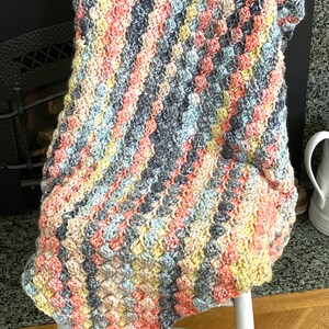 May include: A crocheted blanket with a diagonal pattern in shades of gray, peach, yellow, and blue. The blanket is draped over a white chair.