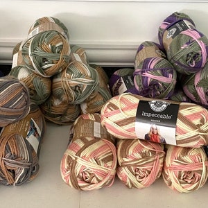 May include: A collection of yarn skeins in various colors and patterns, including brown, beige, pink, green, purple, and blue. The yarn is arranged in stacks and labeled "Impeccable" with a small image of a woman wearing a scarf.
