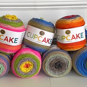 May include: A collection of 14 colorful yarn skeins with the word "CUPCAKE" printed on the label. The yarn is arranged in two rows, with seven skeins in each row. The yarn is a variety of colors, including red, orange, yellow, green, blue, purple, and pink.