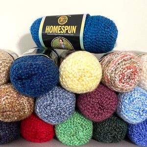 May include: A stack of yarn balls in various colors, including beige, blue, yellow, red, green, and purple. The yarn is arranged in a pyramid shape, with the top ball being a blue yarn ball with a label that reads "HOMESPUN".