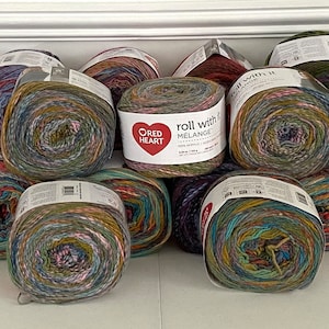 May include: A collection of colorful yarn skeins in various shades of blue, green, brown, and purple. The yarn is wrapped in plastic and labeled "Roll With It" and "Crafty Heart".