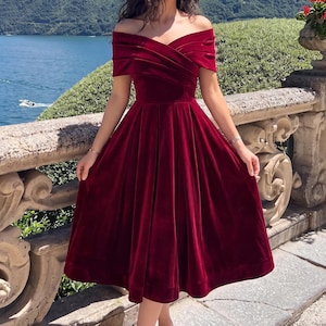 May include: A deep red velvet midi dress featuring an off-the-shoulder neckline with a crossover bodice design. The garment has a fitted waistline that transitions into a pleated, flared skirt that falls below the knees, showcasing a smooth texture.