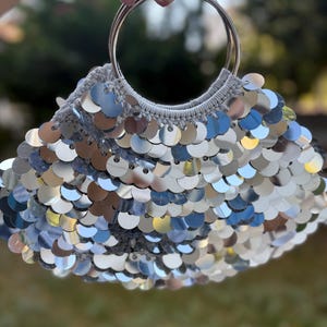 May include: A silver sequin handbag with a crocheted top and two large silver metal handles. The bag is round and covered in shiny, reflective sequins. The background is blurred green.