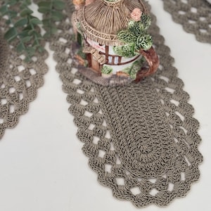 Puede incluir: Primer plano de un camino de mesa de ganchillo gris con forma ovalada y diseño calado. Una casa decorativa de cerámica con techo de paja y detalles de cactus se encuentra sobre el camino de mesa. El camino de mesa está sobre una superficie blanca.