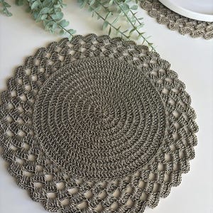 Può includere: Un sottopentola rotondo all'uncinetto grigio e bianco, con un centro a spirale e un bordo smerlato. Il sottopentola è realizzato con un filato strutturato ed è posto su una superficie bianca. Sono visibili anche un piatto bianco e del verde.