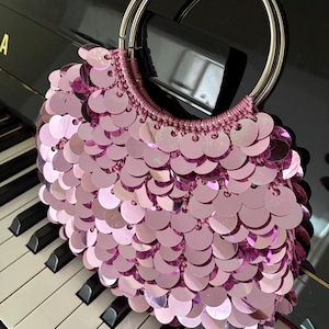 May include: A pink sequin handbag with a silver handle sits on a piano keyboard. The bag is covered in round, shiny sequins. The handle is made of two silver hoops. The piano keys are black and white.