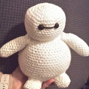 May include: A handmade, white crochet plush toy resembling a character with a rounded body, small arms, and feet. The toy has black eyes and a black line for a mouth. The plush is held in a person's hand.