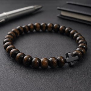 Puede incluir: Una pulsera de cuentas de madera marrón oscuro con un dije de cruz negra. La pulsera es circular y descansa sobre una superficie oscura. Un bolígrafo plateado y un cuaderno negro están en el fondo.